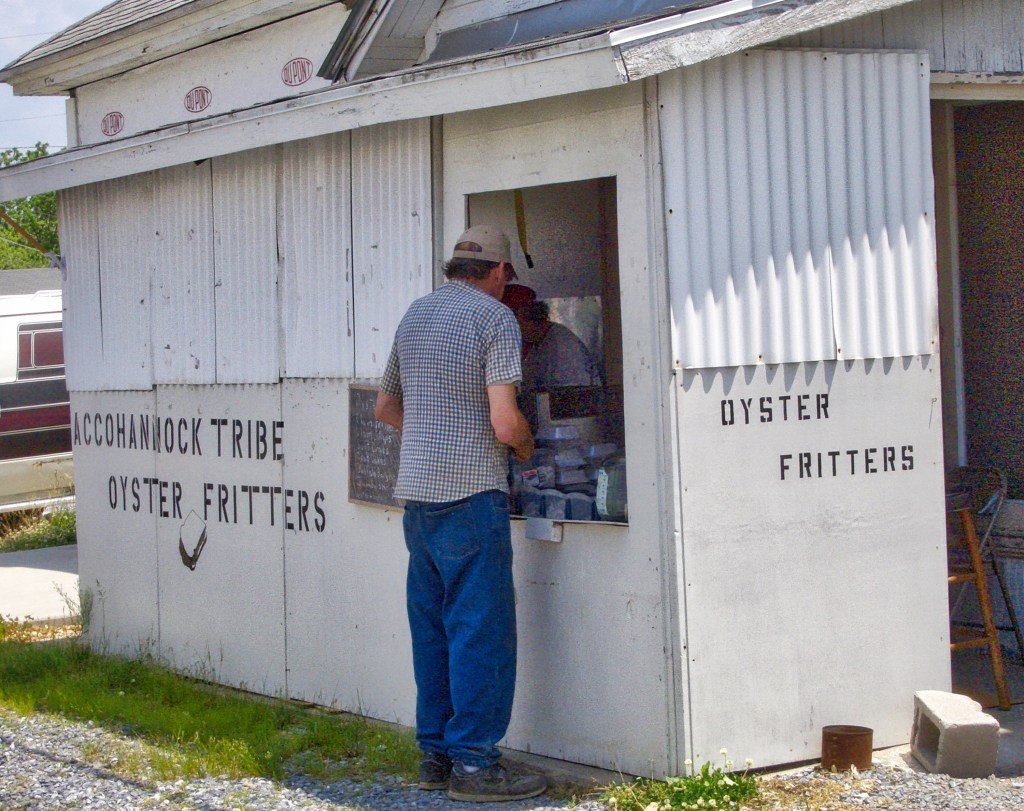 Color photo of a small side building half covered in corrugated panels. Stenciled on the front is ACCOHANNOCK TRIBE OYSTER FRITTERS. A man is standing at the carryout window.