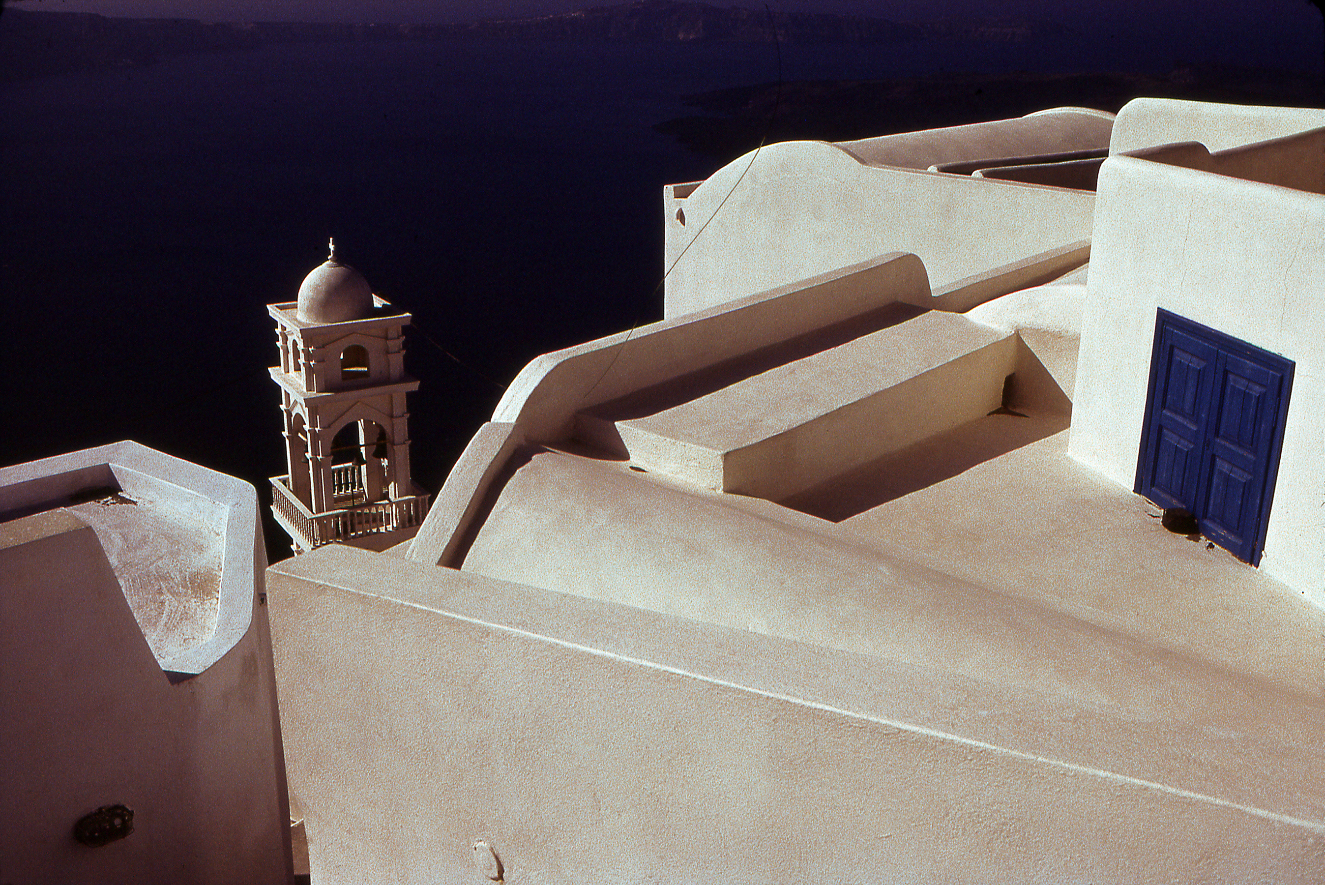 "Church Tower" This is a tall tower with a bell seen in the distance over the tops of the roofs of a small greek village. A pair of blue doors are visible on one of the roofs.